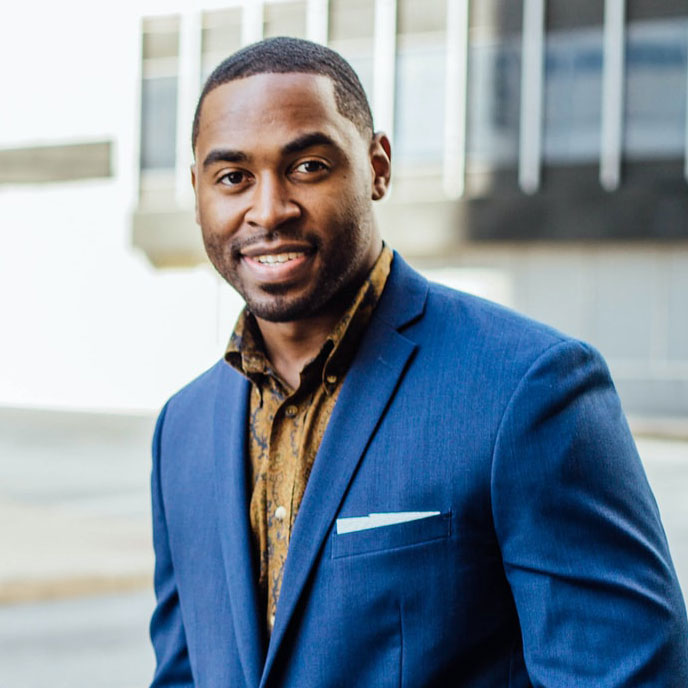 Smiling man wearing a dark blue suit jacket and a patterned brown shirt, standing outdoors in an urban setting.