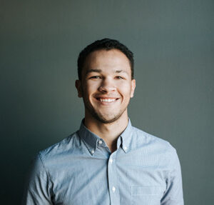 Smiling man wearing a light blue button-up shirt, standing against a plain gray background. He has short curly hair and a friendly expression, facing the camera directly.