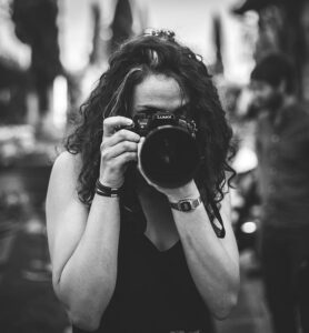 Black and white photo of a woman with curly hair holding a Lumix GH4 camera up to her face, taking a photo.