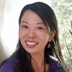 Linling smiling warmly, wearing a purple top and beaded necklace, with long dark hair and star-shaped earrings, sitting near a sunlit window with green foliage in the background.